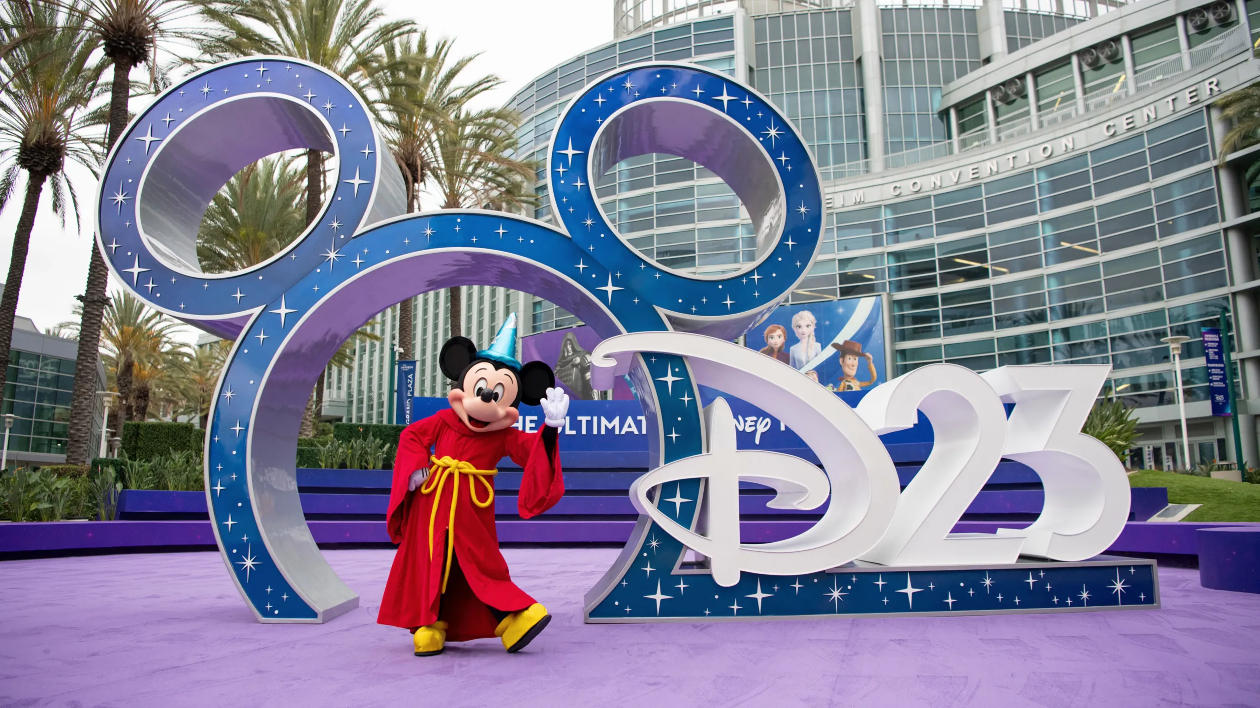 Mickey Mouse in a red sorcerer's robe stands in front of a large Disney D23 display outside the Anaheim Convention Center.