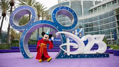 Mickey Mouse in a red sorcerer's robe stands in front of a large Disney D23 display outside the Anaheim Convention Center.