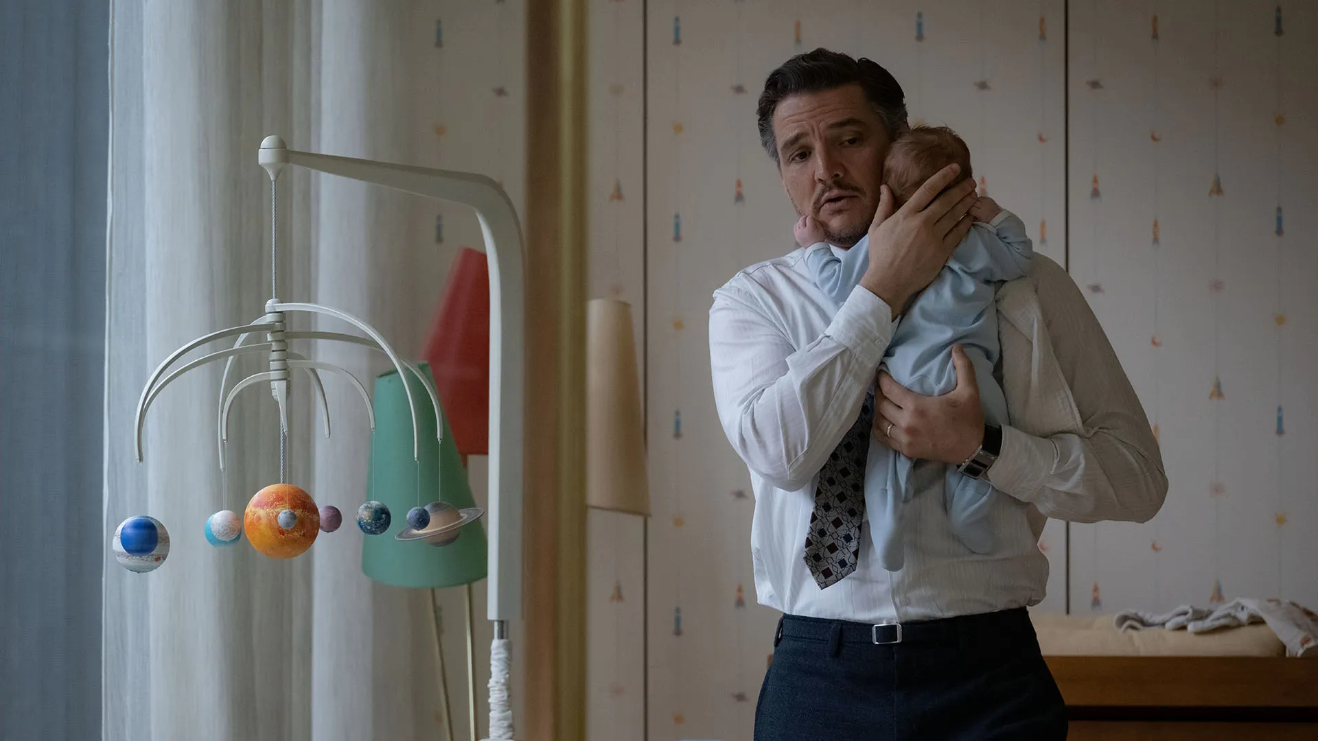 Una escena cinematográfica de Los 4 fantásticos: Primeros pasos muestra a Reed Richards (Pedro Pascal) con expresión preocupada mientras acuna con delicadeza a un bebé en sus brazos, con un colorido móvil para bebés del sistema solar colgando en primer plano