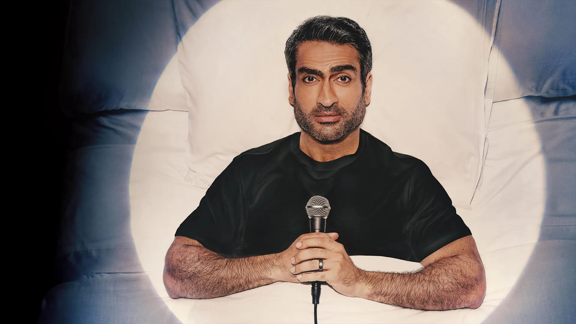 Kumail Nanjiani lying in bed holding a microphone, with a spotlight shining on him