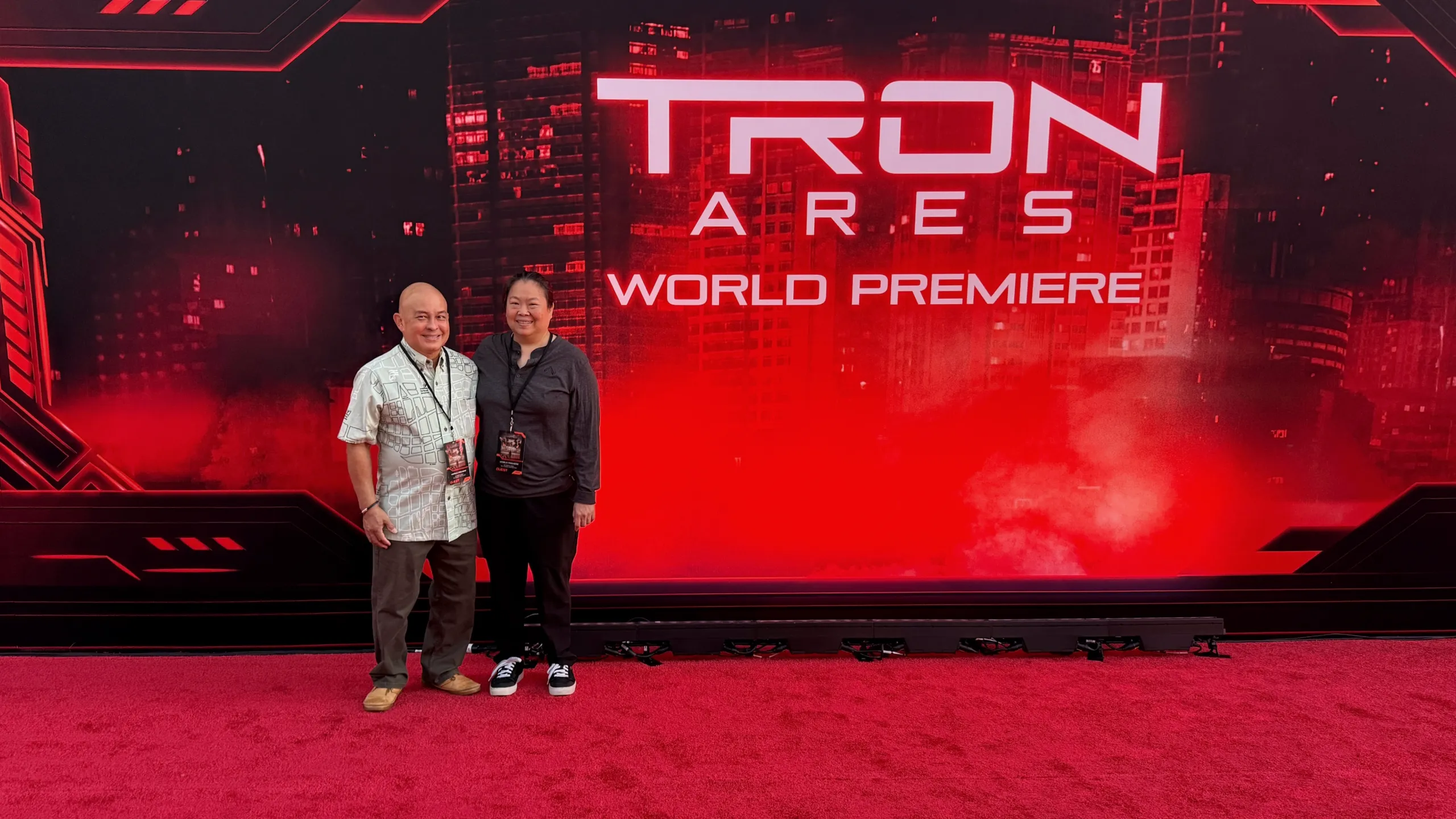 Jeanine C. and her husband standing on the red carpet. The background reads "Tron: Ares World Premiere" superimposed on a red-tinted cityscape.