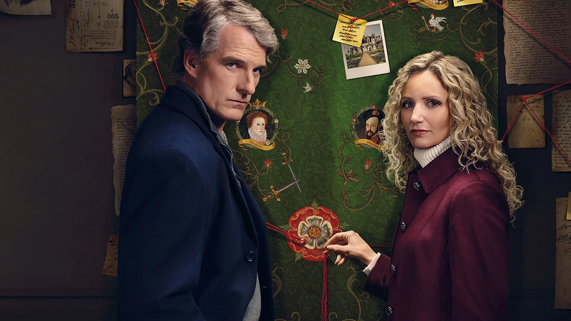 Two people stand beside a large decorative tapestry covered with photographs, notes and red string connecting clues.