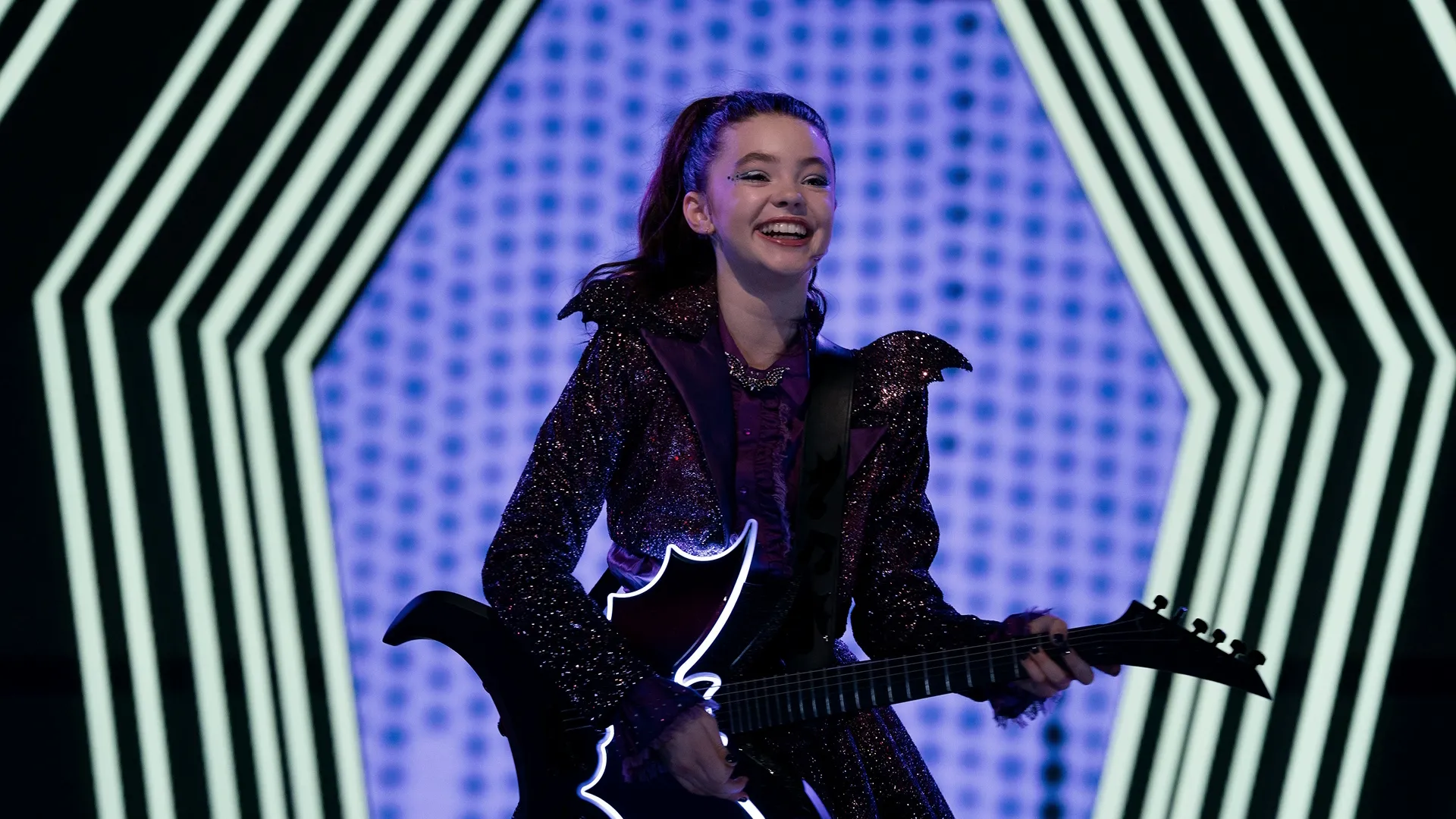 Vee Hauntley (Kenzi Richardson) flashes a wide, enthusiastic smile while playing a black electric guitar on a brightly lit stage in front of a blue and white geometric backdrop.