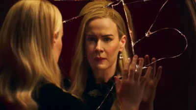 Cordelia Goode (Sarah Paulson) olha fixamente para um espelho circular muito rachado com uma expressão aflita. A mão dela repousa suavemente sobre o vidro quebrado, enquanto seu reflexo reproduz seu olhar preocupado. Ela usa uma blusa preta com babados adornada com um broche vintage, além de brincos de cristal pendentes que contrastam com o fundo vermelho quente e mal iluminado.