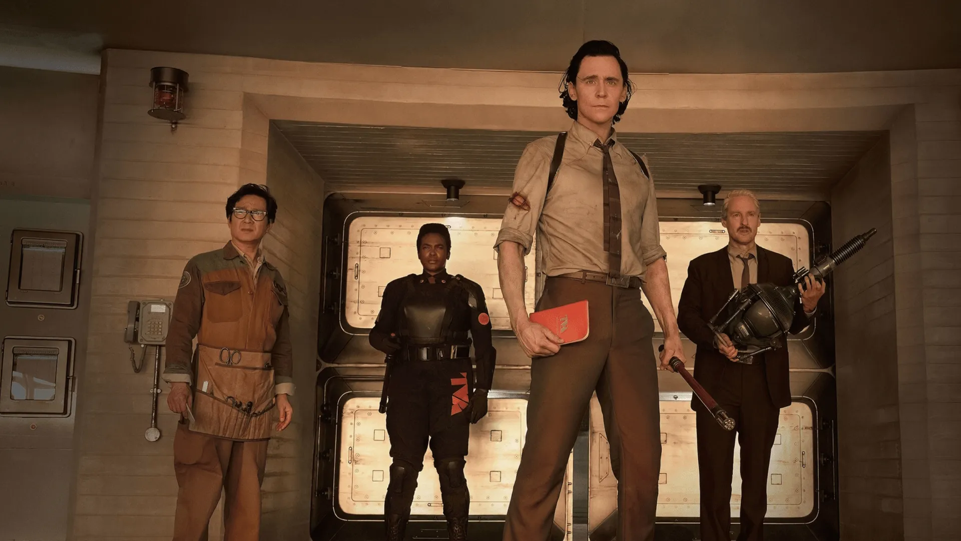 Tom Hiddleston as Loki holding a handbook, standing alongside Owen Wilson, Ke Huy Quan, and Wunmi Mosaku inside the TVA headquarters in a scene from Marvel Studio's Disney Plus series Loki Season 2.