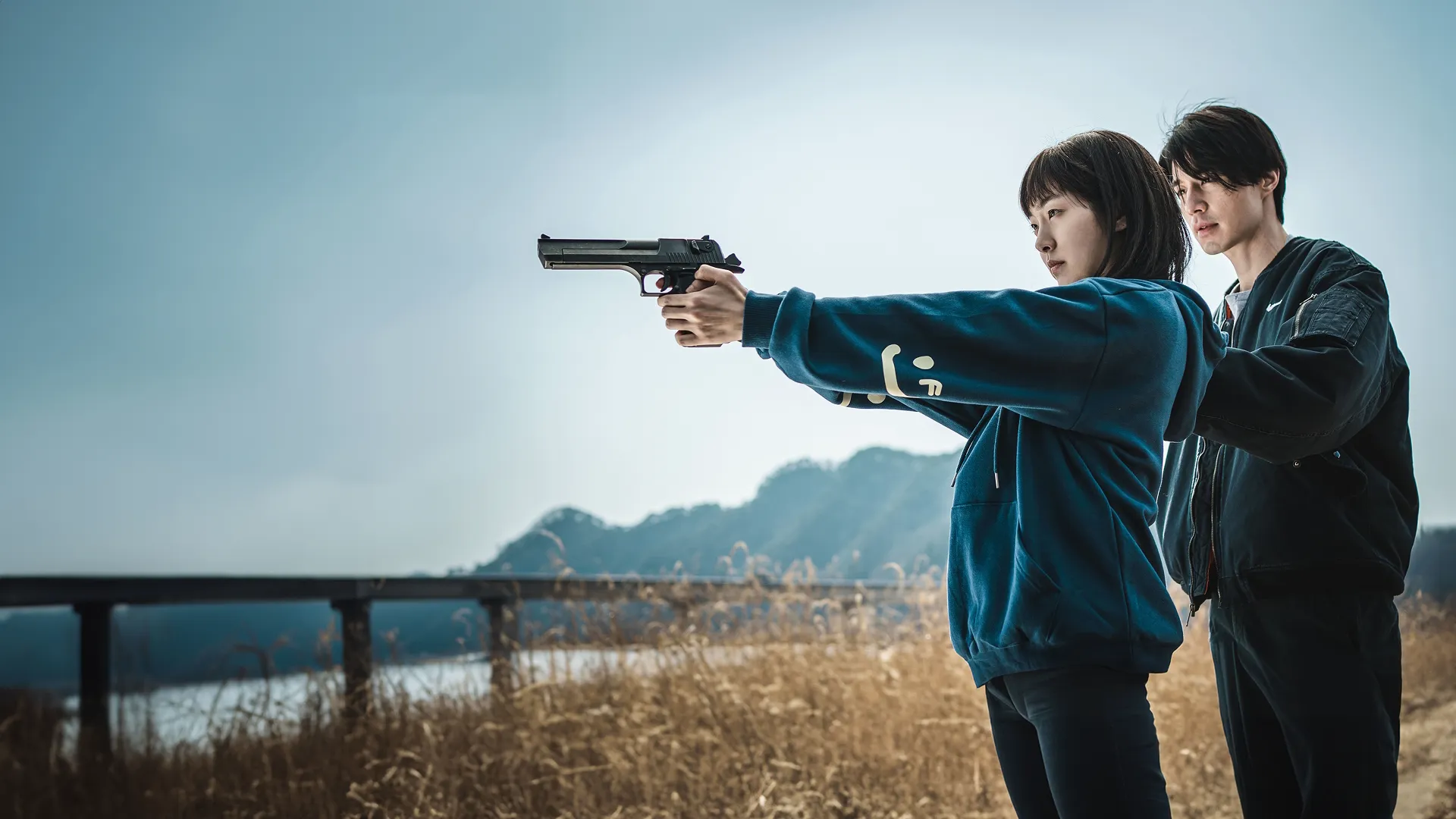 A dramatic promotional image from "A Shop for Killers" showing a young woman in a blue hoodie holding a large pistol with both hands, aiming it forward with focus, while a man in a black jacket stands closely behind her, guiding her posture against an outdoor background of dry grass, hills, and an overcast sky.