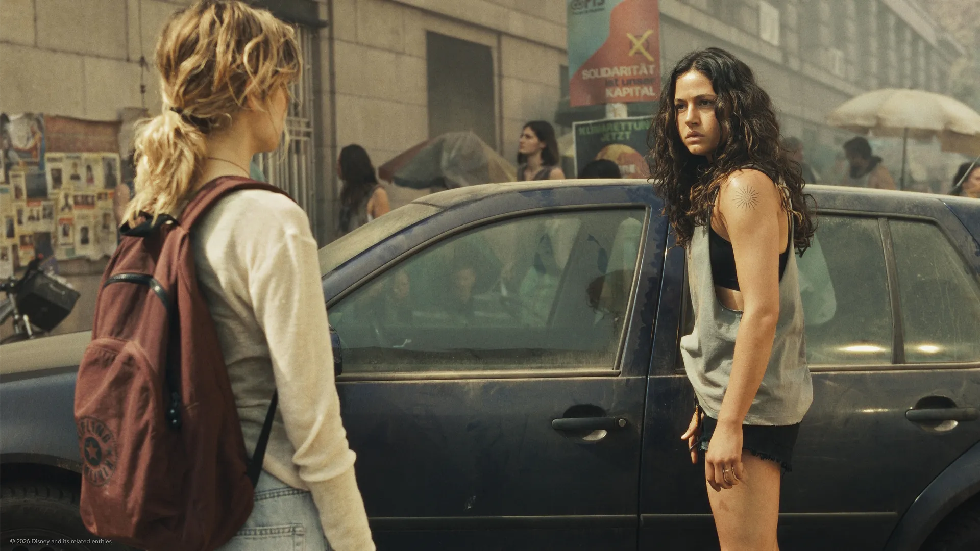 An official scene from the dystopian drama series City of Blood, featuring a tense standoff between two young women standing on a dusty city street next to a dark car.