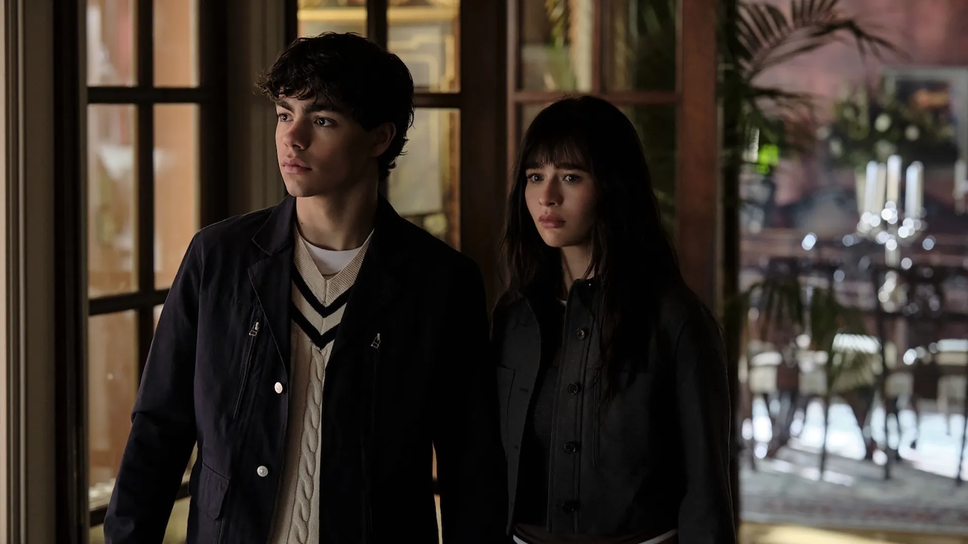 In a scene from the supernatural series Coven Academy, Jake (Malachi Barton) and Briar (Malina Weissman) look off to the side with anxious, worried expressions while standing near a large glass pane window indoors.