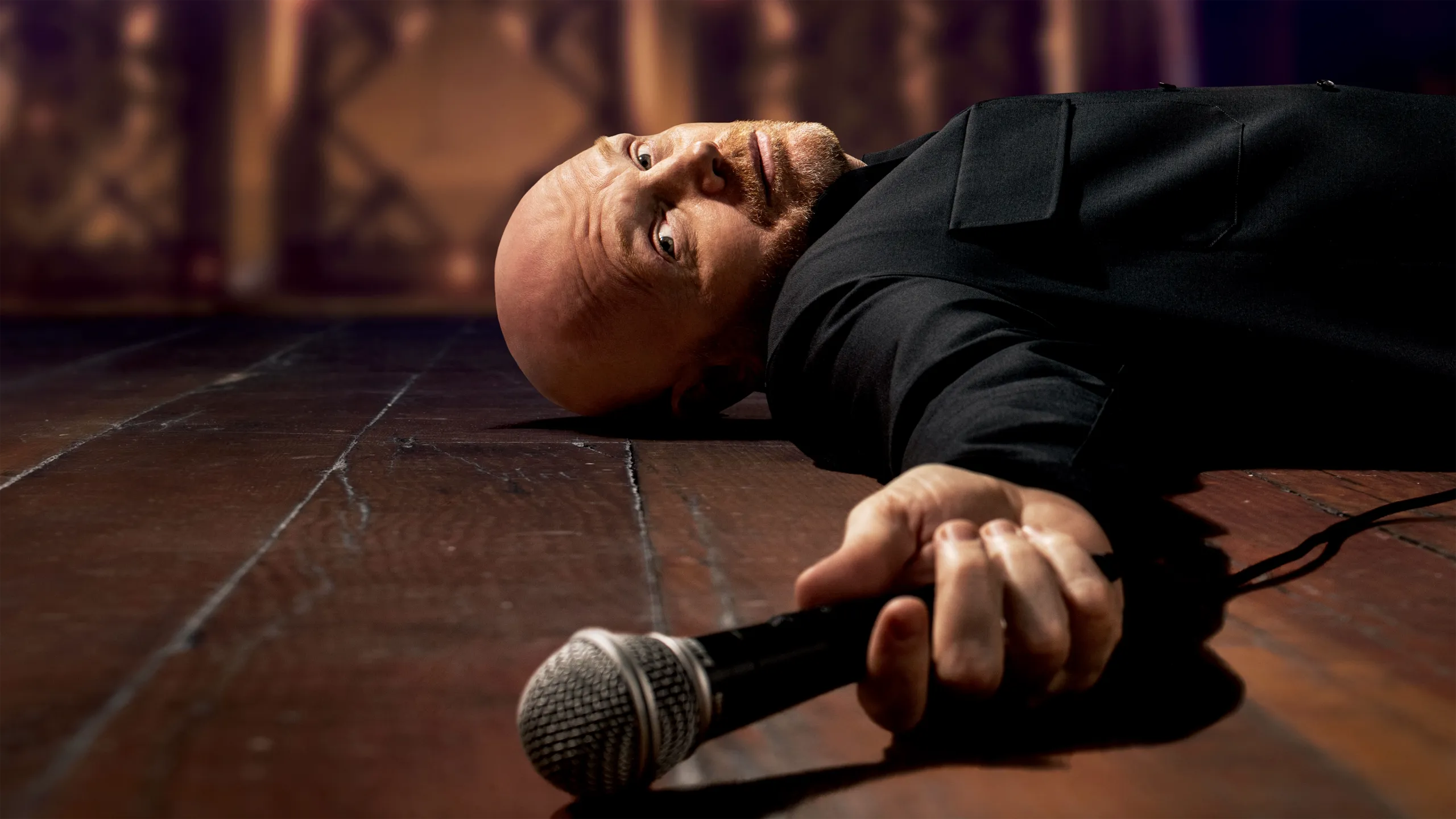The image shows comedian Bill Burr lying on a stage floor, holding a microphone. 