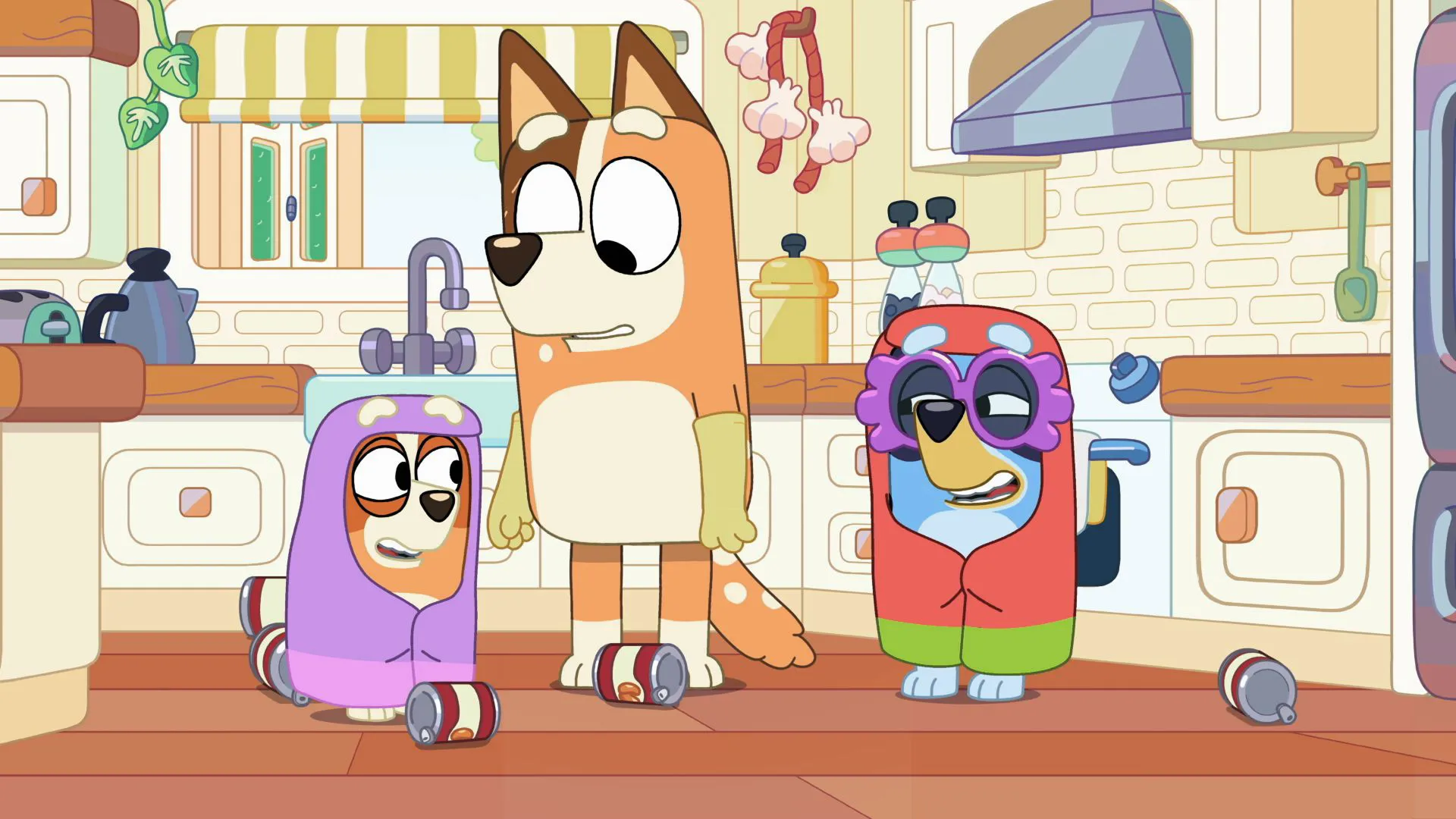 Chilli stands in the kitchen looking surprised as Bluey and Bingo, dressed up as the "Grannies" Janet and Rita, cause a mess with soup cans on the floor.