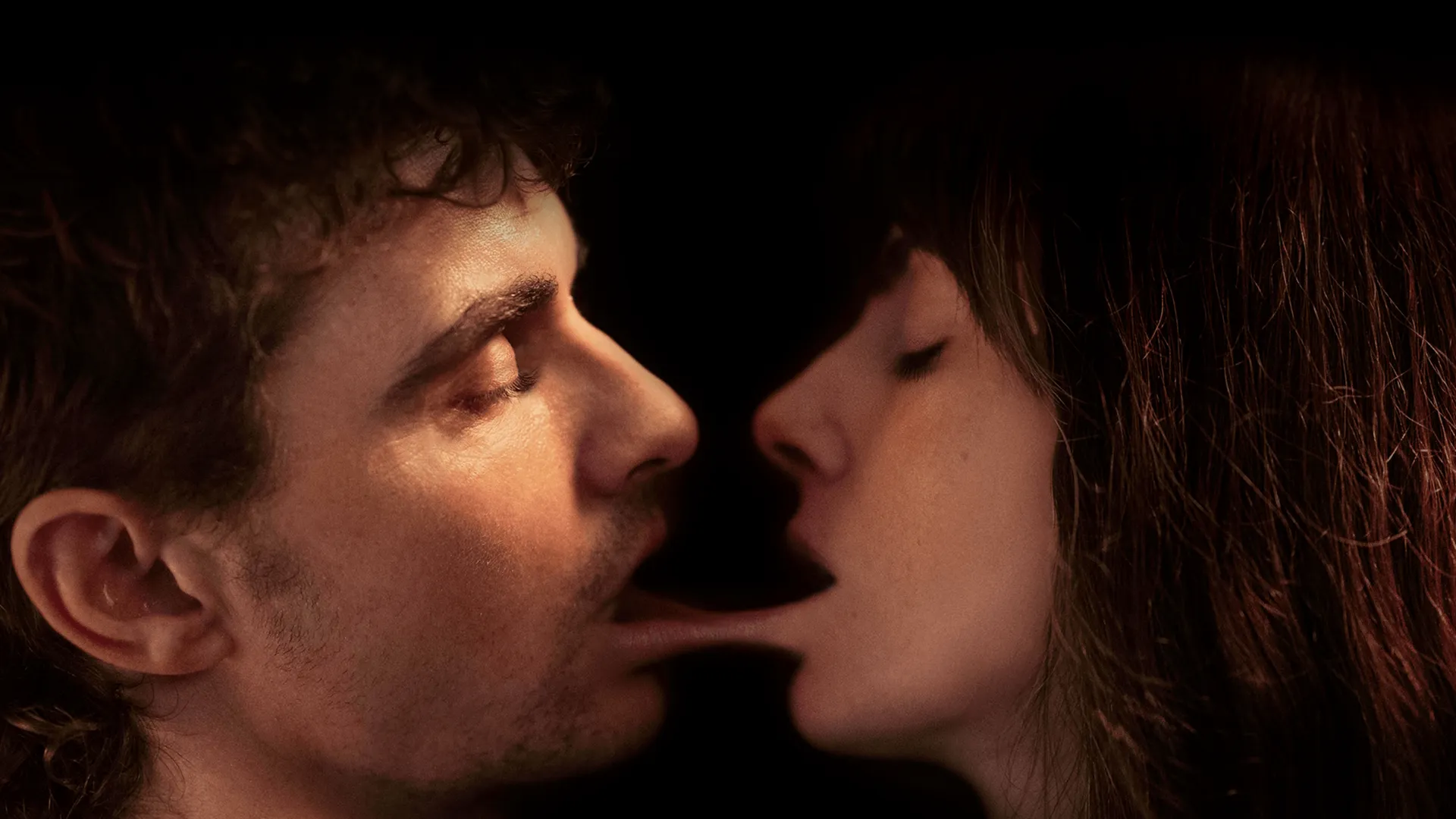 A striking, intimate close-up film still from the supernatural body horror movie Together, showing Tim (Dave Franco) and Millie (Alison Brie) facing each other in profile with their eyes closed as a surreal, stretched layer of flesh connects their open mouths against a black background.