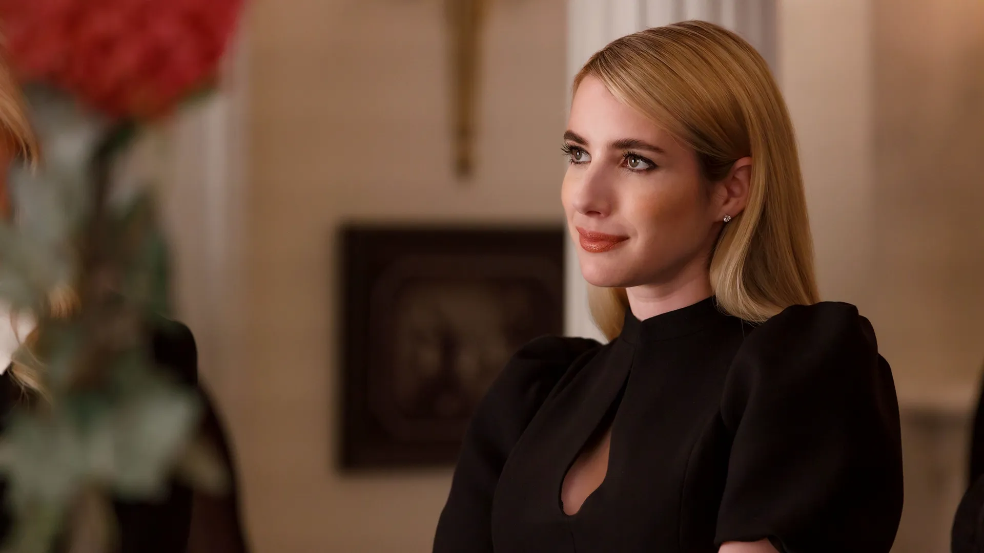 A blonde woman with her hair sleekly parted to the side looks off with a subtle, knowing smile, wearing a black dress featuring puffed sleeves and a small keyhole cutout at the neckline.