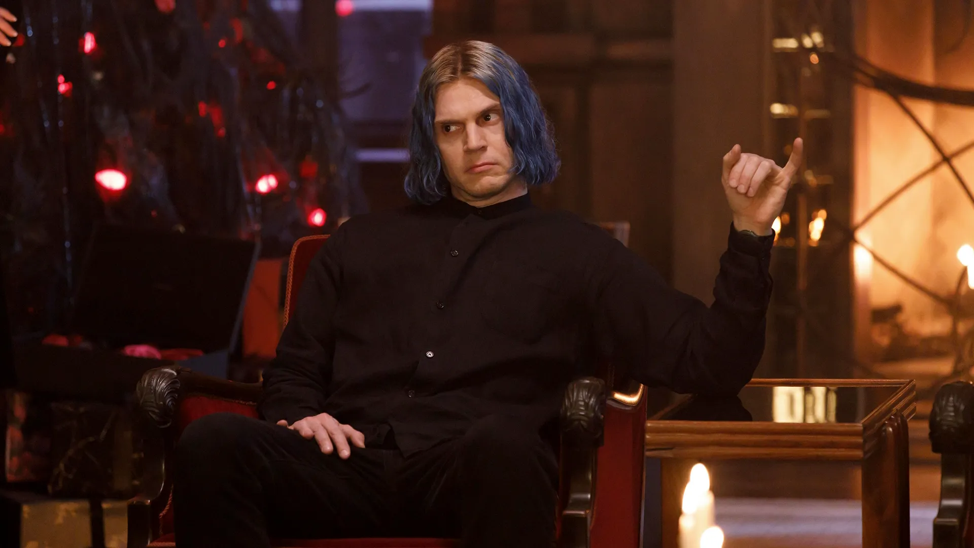 A man with chin-length blue hair and dark roots sits in an armchair wearing an all-black button-down shirt, making a skeptical expression while raising one hand with a curled pinky finger.