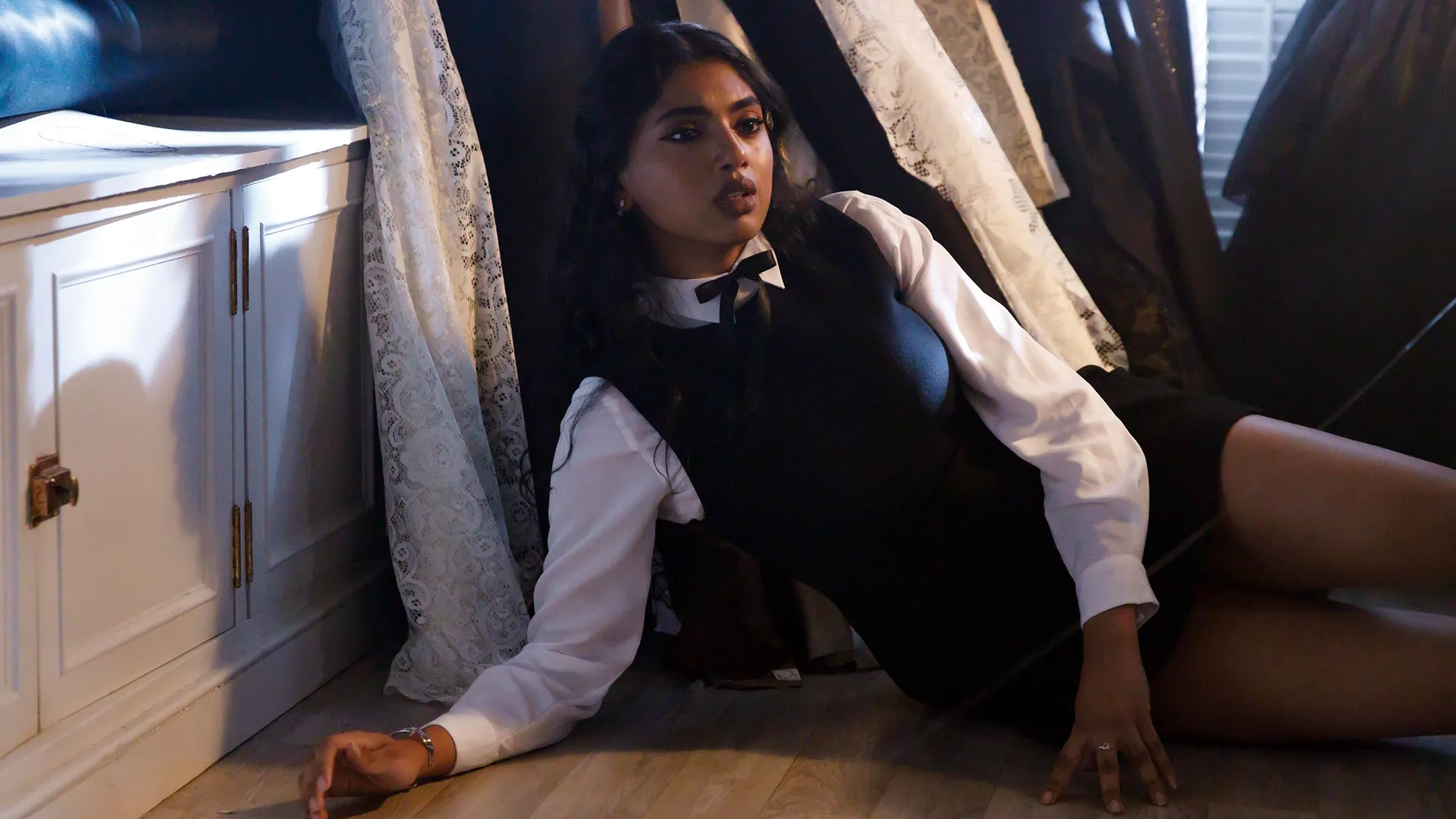 A young woman with long dark hair lies on a wooden floor leaning against a white cabinet, wearing a black pinafore dress over a long-sleeved white collared shirt with a black ribbon bow tie.