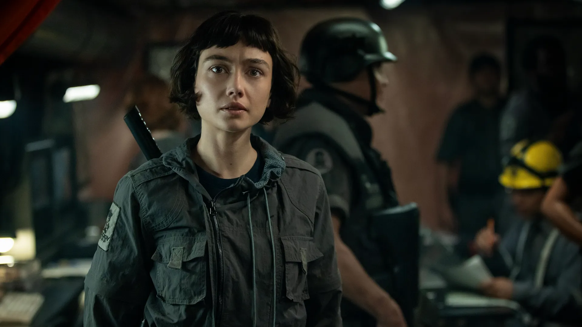 Wendy (Sydney Chandler) looking straight ahead with wide, intense eyes, dressed in a grey tactical zippered jacket with a shoulder patch, while a soldier in a helmet stands out of focus behind her in a dimly lit control room.