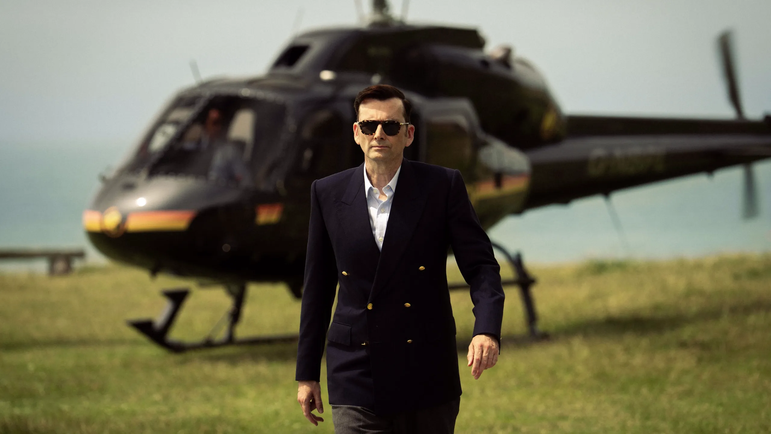 Tony Baddingham (David Tennant) walks across a grassy field with a helicopter behind in Rivals season 2.