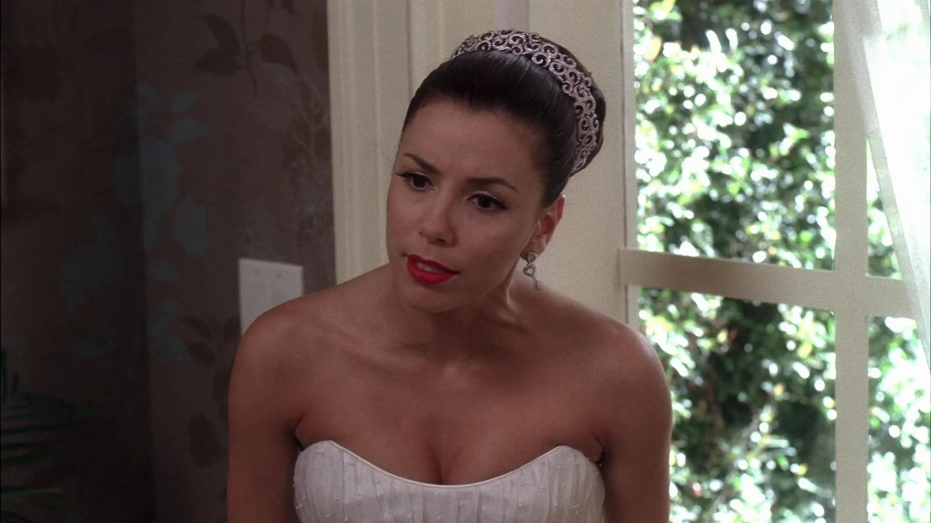 Gabrielle Solis (Eva Longoria) looks forward with a concerned expression, wearing a strapless white gown, a jeweled tiara over her updo, and bright red lipstick next to a bright window.