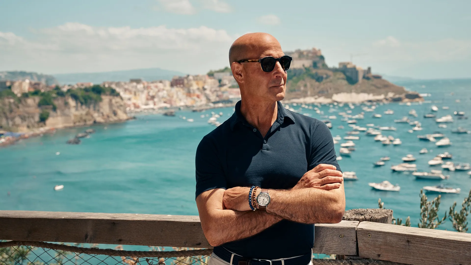 Stanley Tucci de pie frente a un paisaje costero italiano, con los brazos cruzados y anteojos de sol