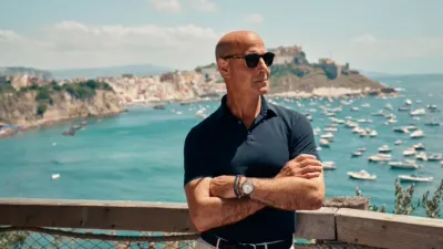 Stanley Tucci de pie frente a un paisaje costero italiano, con los brazos cruzados y anteojos de sol