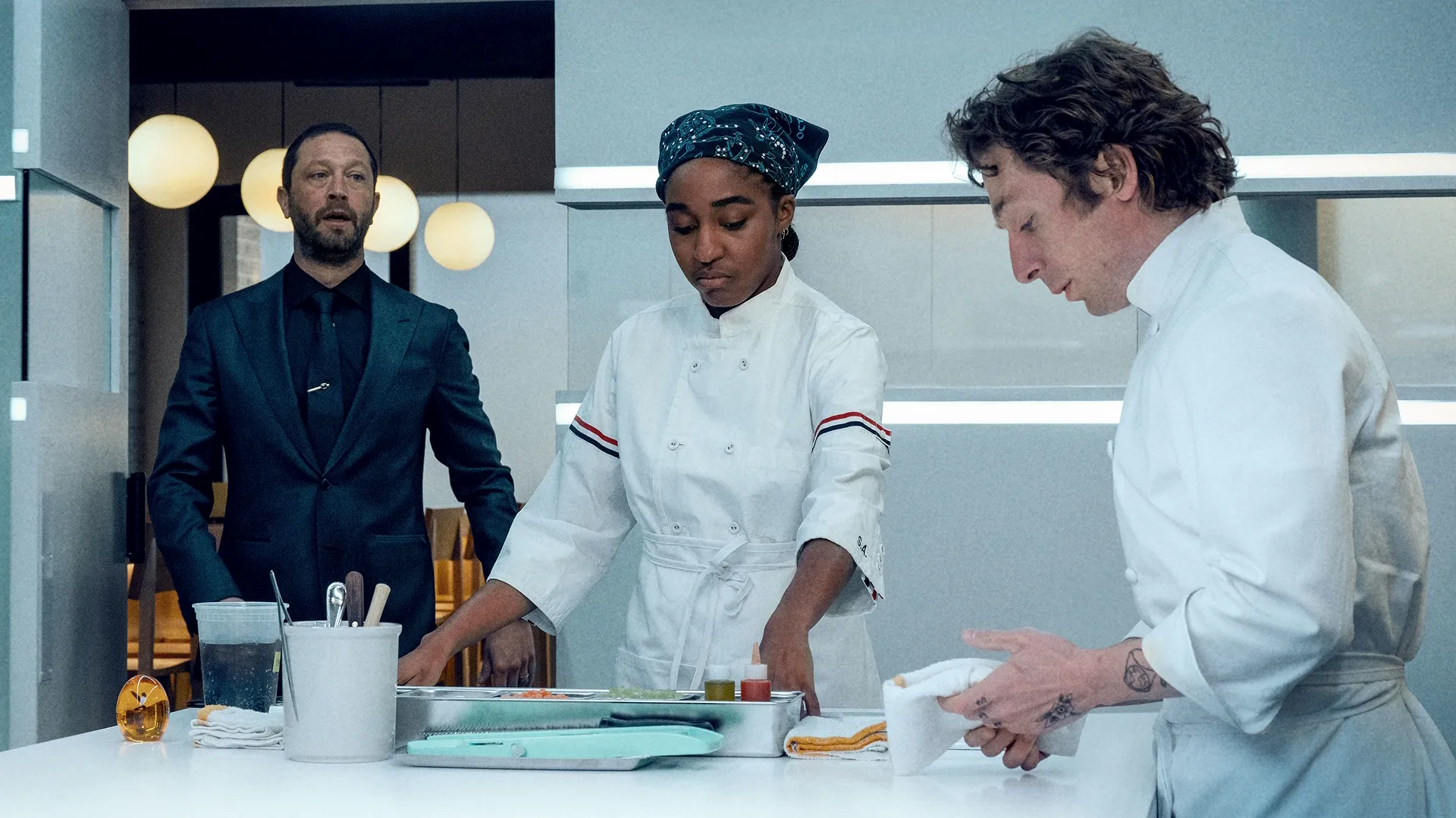 Richie (Ebon Moss-Bachrach) looks on while Sydney (Ayo Edebiri) and Carmy (Jeremy Allen White) prepare food in a kitchen