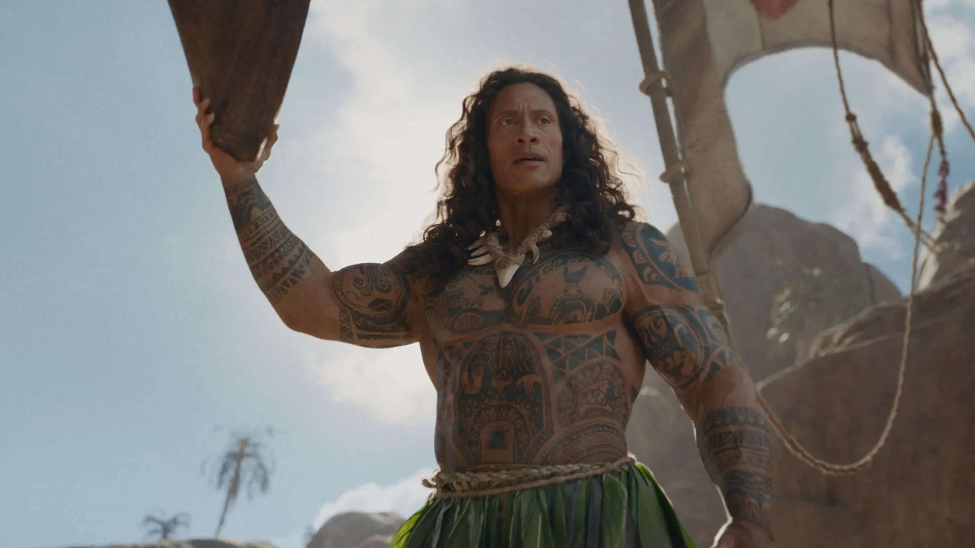 Dwayne Johnson as a live-action Maui stands shirtless with a serious expression, displaying his extensive tribal tattoos and wearing a leaf skirt under a bright, partly cloudy sky.