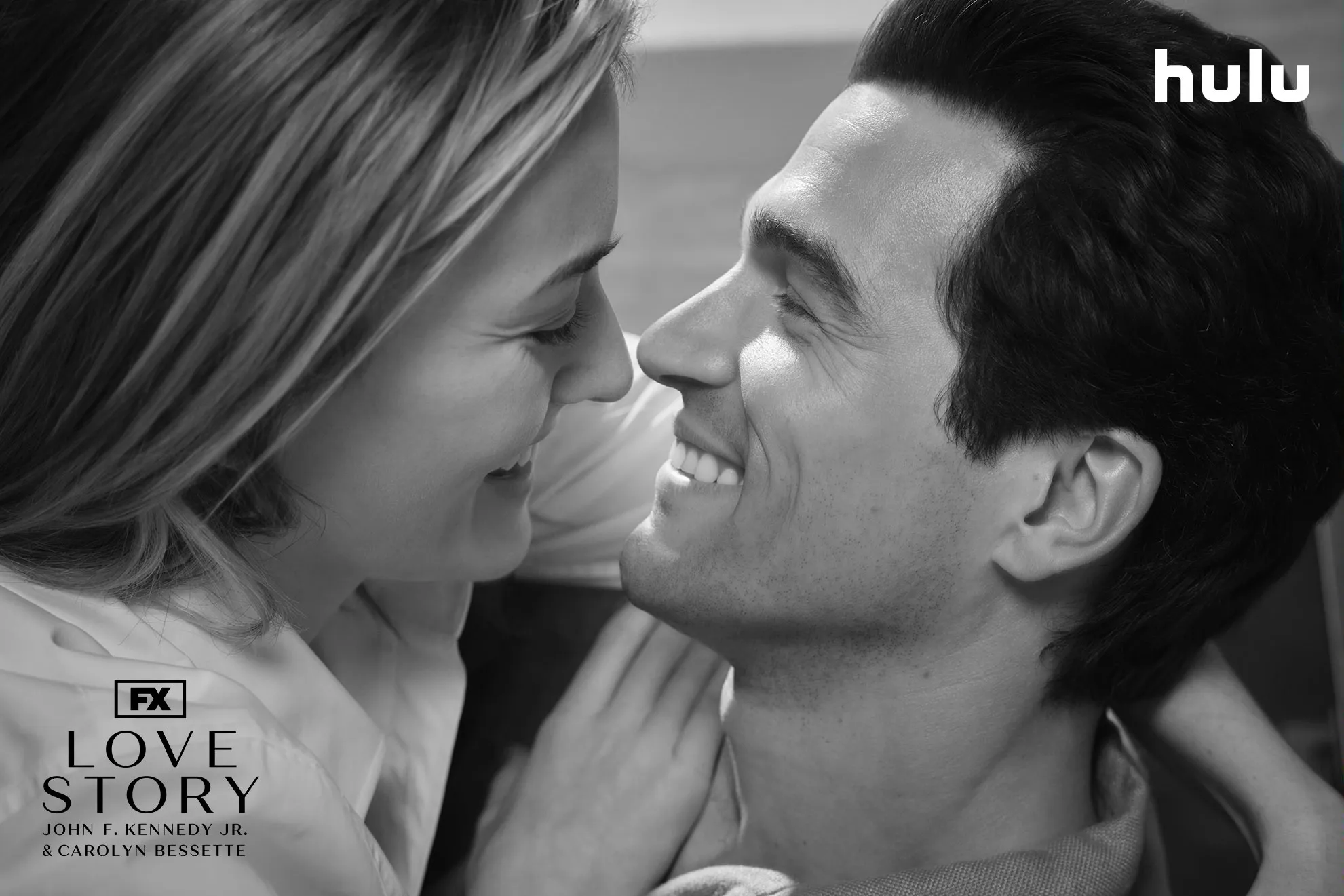 Close-up of Carolyn Bessette (Sarah Pidgeon) and John F. Kennedy Jr. (Paul Kelly) embracing in a black and white set photo from FX's Love Story.