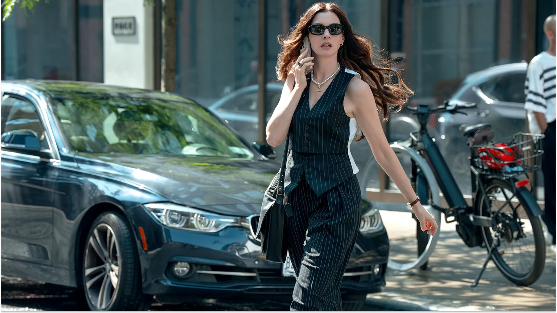 A promotional still from The Devil Wears Prada 2 captures Andy Sachs (Anne Hathaway) hastily crossing a New York street in a chic vintage Jean Paul Gaultier pinstripe vest and matching trousers. She is wearing dark sunglasses, layered necklaces, and talking into a smartphone held to her ear as she walks past a parked dark sedan.
