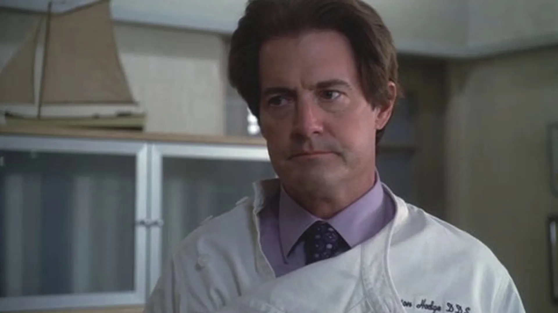 Orson Hodge (Kyle MacLachlan) stands in his dental office with a tight, tense expression, wearing a purple dress shirt, a patterned tie, and a white dentist's smock embroidered with his name.