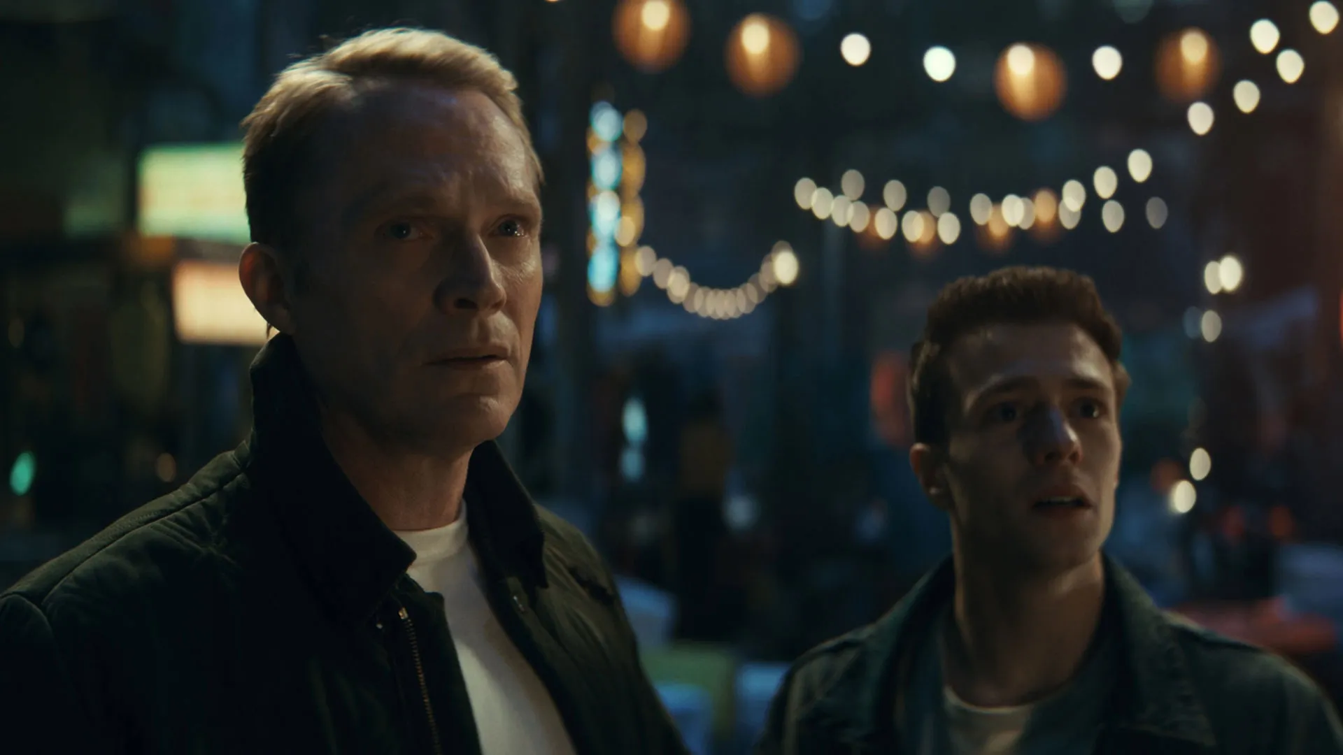 Vision and Tommy Maximoff stand side-by-side with expressions of concern and surprise against a blurred backdrop of string lights in Marvel's VisionQuest.