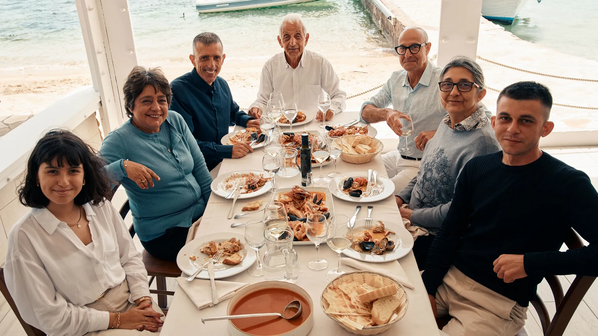 Stanley Tucci sits with a family at a dining table after a meal, with a variety of dishes and plates set out