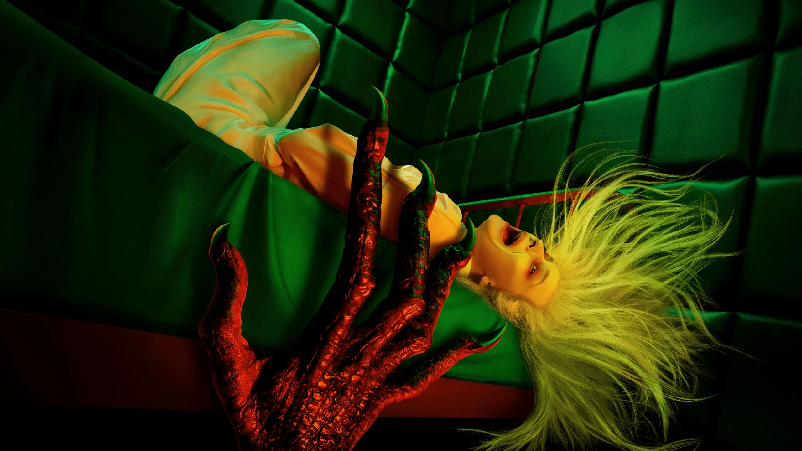 A striking, horror-themed image featuring a woman with long blonde hair screaming while upside down on a bed, as a monstrous, clawed red hand reaches up into the foreground against a backdrop of green, padded walls.