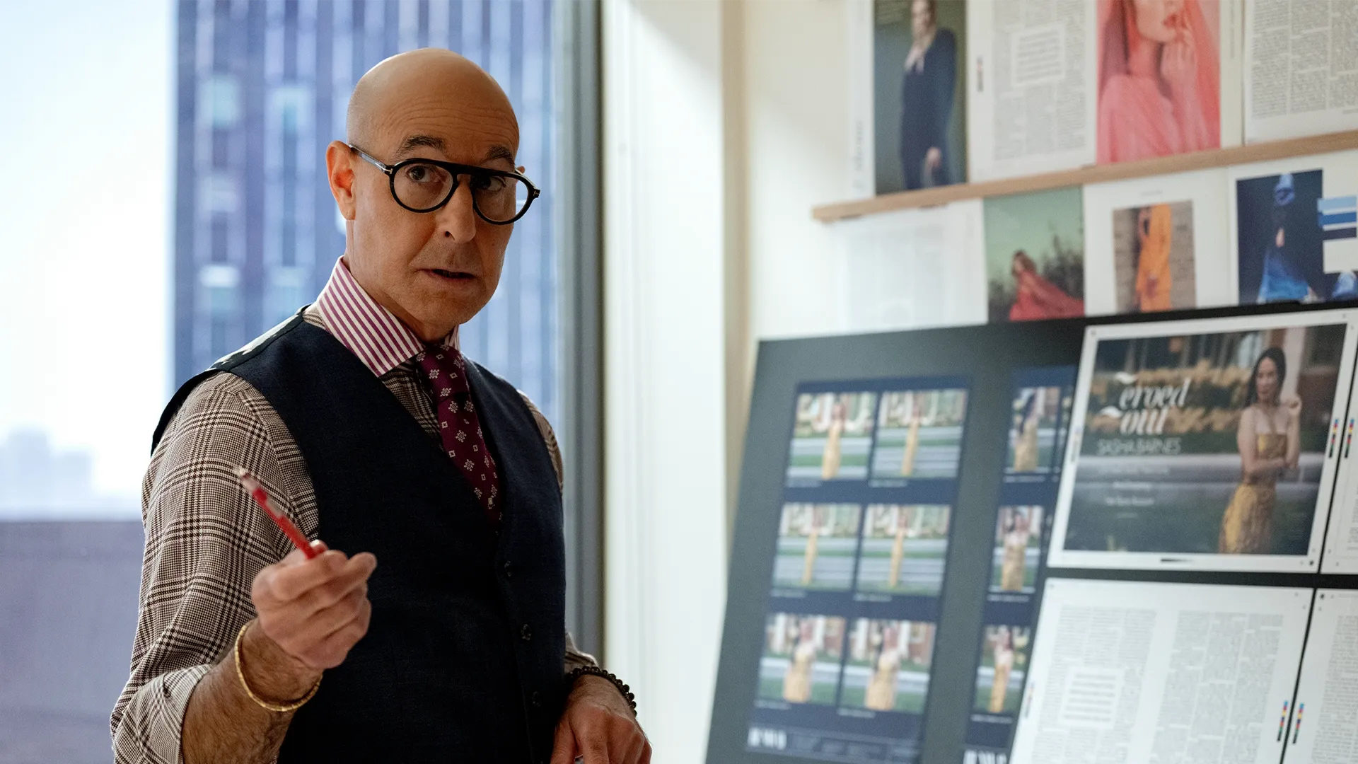 A promotional still from The Devil Wears Prada 2 shows Nigel Kipling (Stanley Tucci) standing in a brightly lit fashion office, holding a red pencil and gesturing while speaking with a surprised expression. He wears thick round black glasses, a navy waistcoat over a brown houndstooth shirt, a red-and-white striped collar, and a patterned purple tie, with fashion layout boards visible behind him.