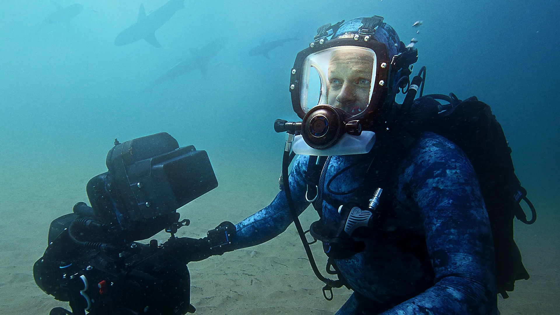 Bertie Gregory under the water in scuba gear and a camera with sharks visible in the distance behind him.