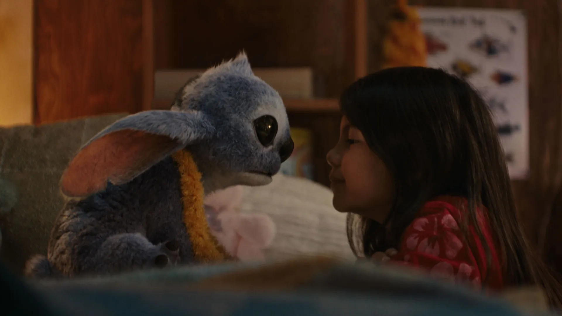 Stitch, wearing a yellow flower lei, sits face to face with Lilo on her bed.
