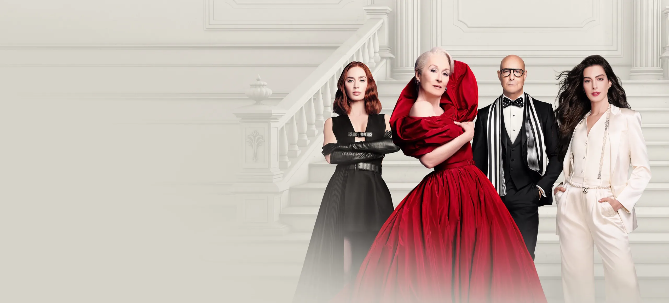 Four actors from the movie The Devil Wears Prada—Emily Blunt, Meryl Streep, Stanley Tucci, and Anne Hathaway—pose together in high-fashion black, red, and white outfits on a grand marble staircase.