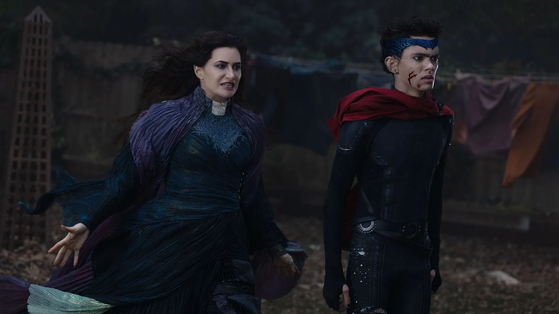 Agatha Harkness and Teen wearing his Wiccan costume stand side by side in a dimly lit, outdoor setting from Marvel's Agatha All Along.