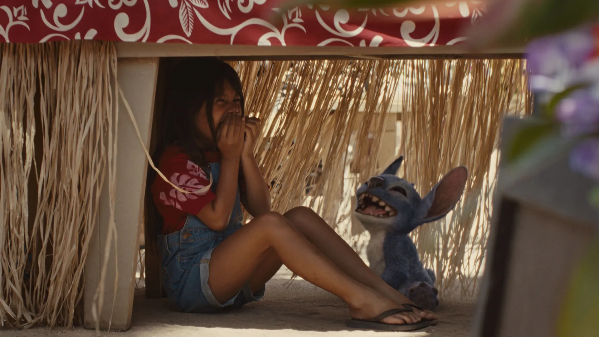Lilo and Stitch laugh together while hiding under a table on the beach.