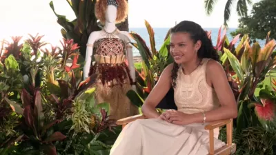 Catherine Laga'aia wearing a cream dress and being interviewed by a tropical beach