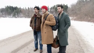 The Jonas Brothers stand in the middle of a country road covered in slush with fresh snow at the sides of the road.