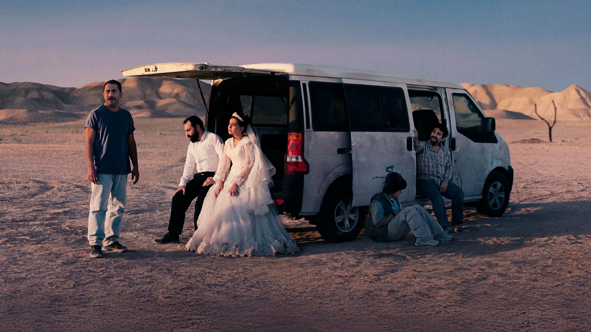 A bride and four men rest by a white mini-van in a vast desert landscape.