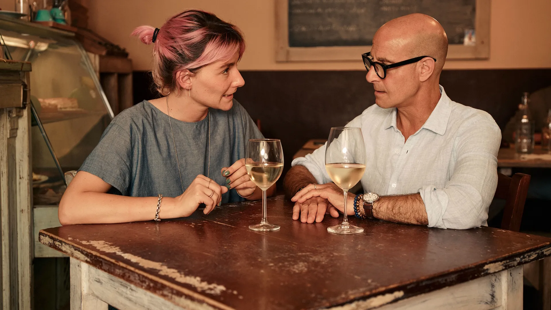 Stanley Tucci sits with a woman at a table with two glasses of white wine.