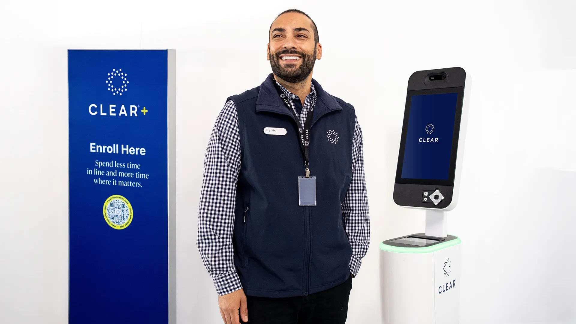 Man in professional attire standing with a Clear+ billboard and a smartphone with the Clear+ app on-screen.