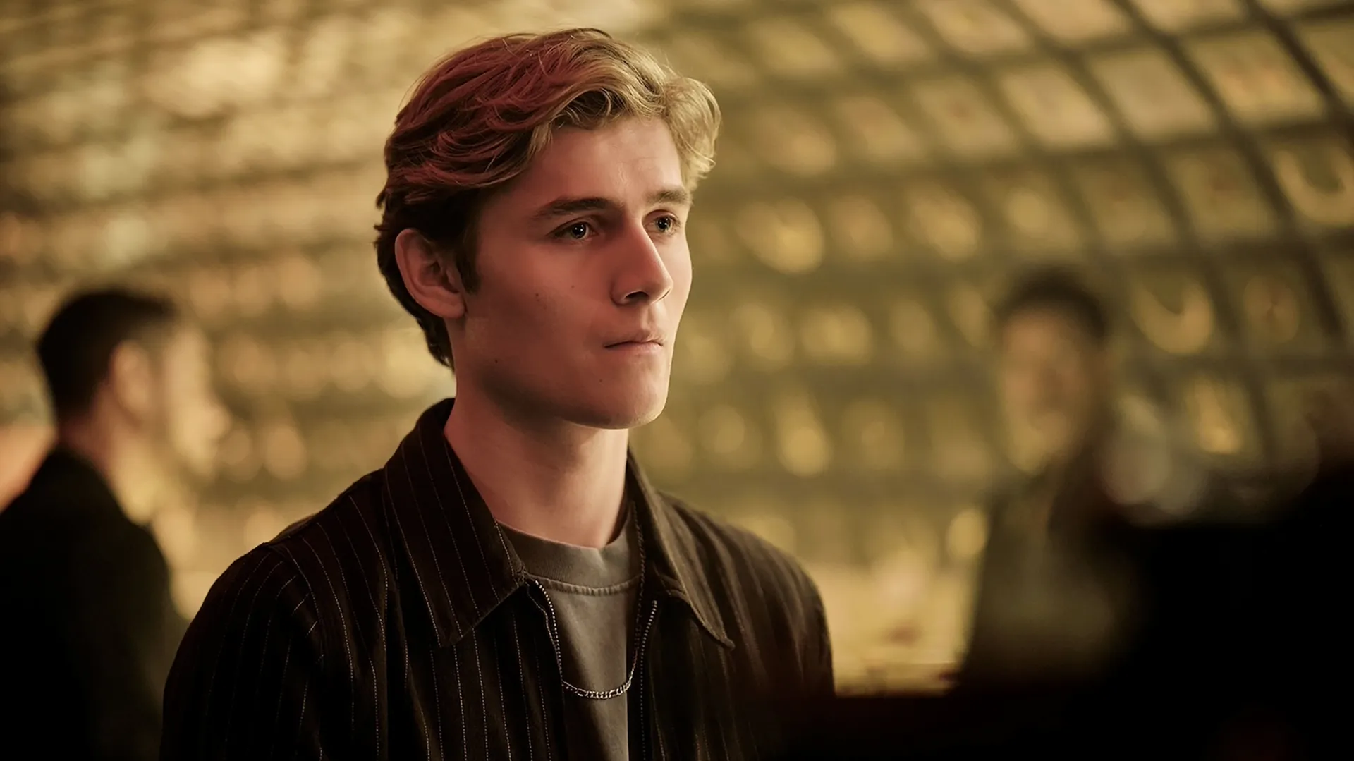 In a scene from the supernatural series Coven Academy, Ollie (Louis Thresher) looks ahead thoughtfully with a subtle pout, wearing a dark pinstripe jacket over a grey shirt and a silver chain necklace.