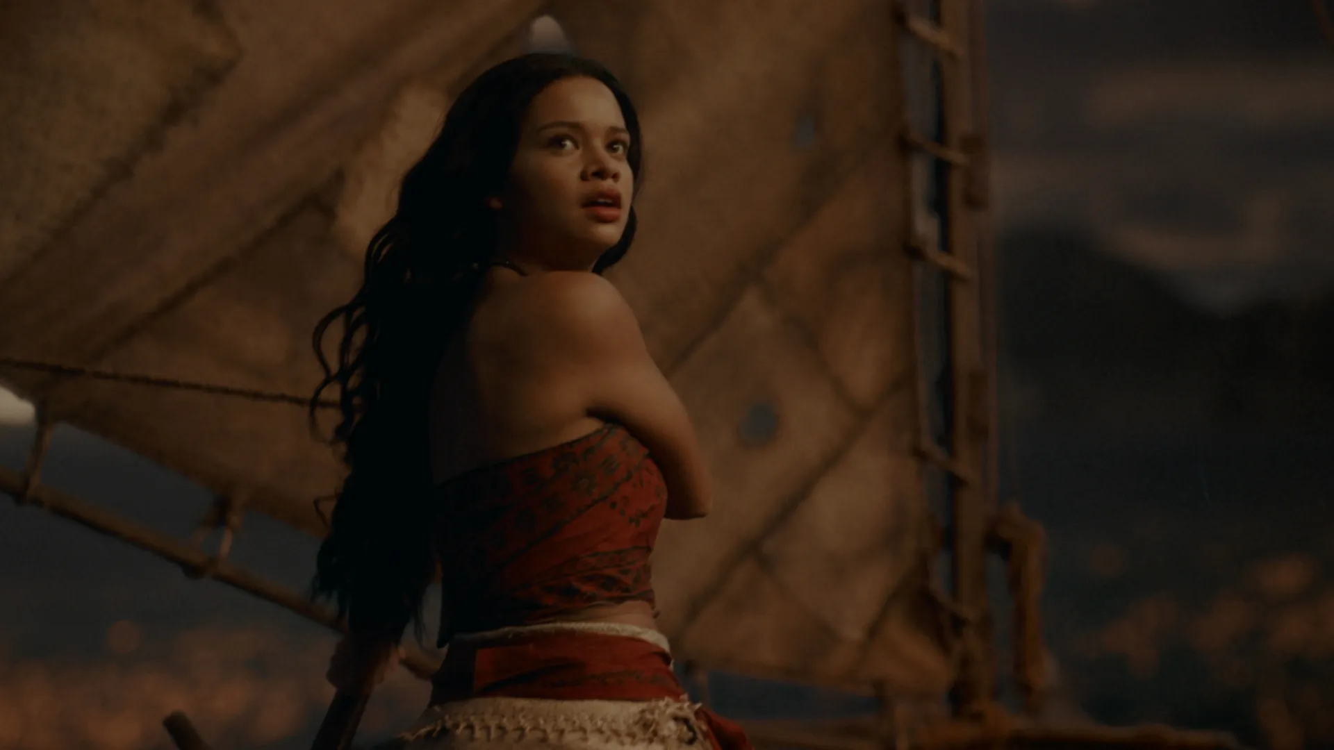 A live-action Moana stands on a sailboat, looking over her shoulder with an alarmed expression against a backdrop of sails and a dim, warm-toned horizon.