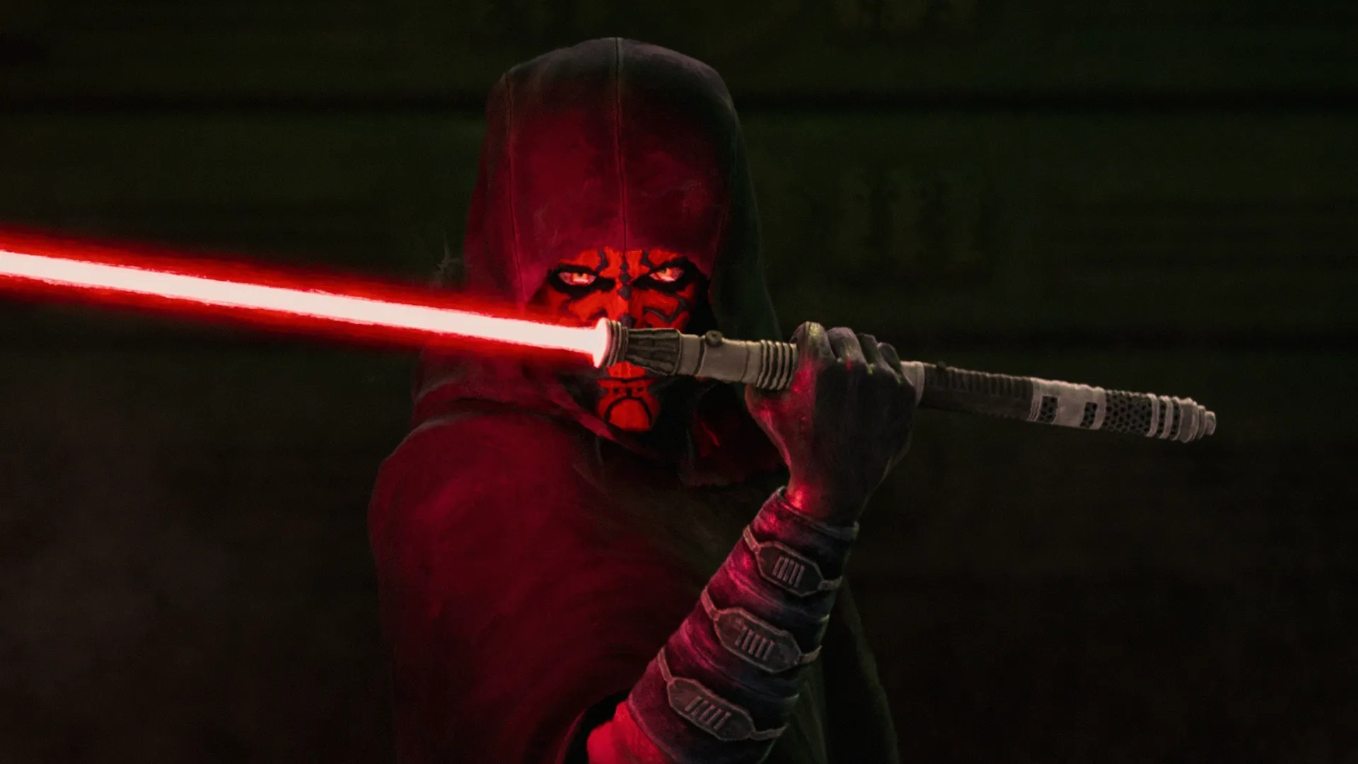 A cloaked Maul in a dark are holds his red lightsaber which illuminates his face.