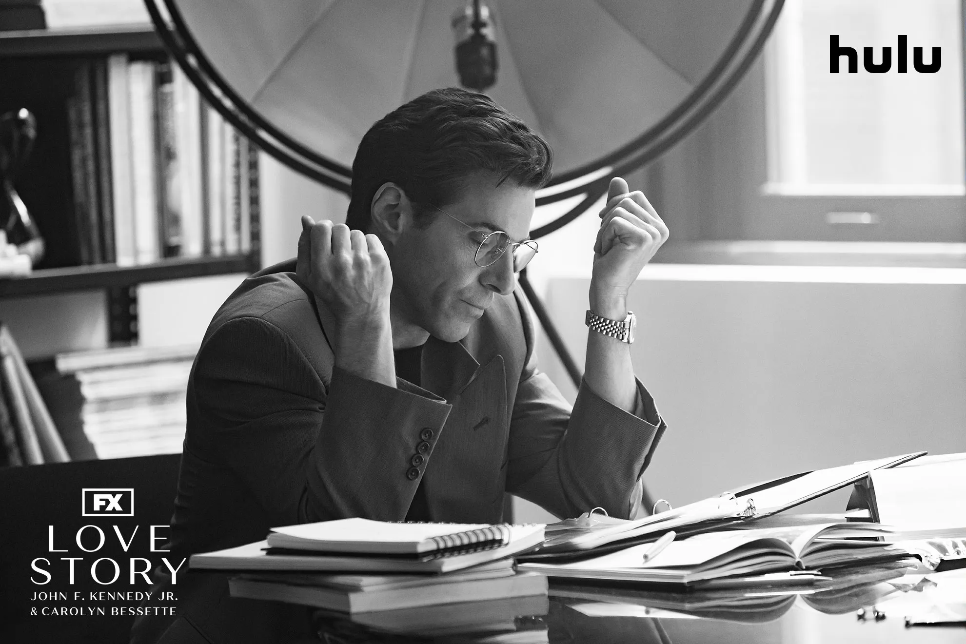 A focused John F. Kennedy Jr. (Paul Kelly) at his desk in a black and white set photo from FX's Love Story.