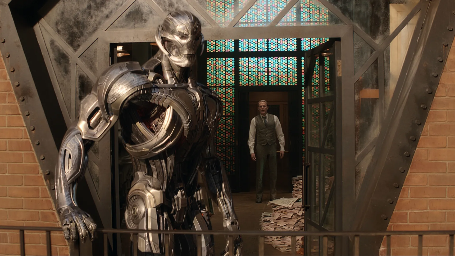 The metallic, robotic form of Ultron stands menacingly in the foreground outside a building, while White Vision in human disguise looks out at him from a doorway behind stacks of old newspapers in Marvel Television's VisionQuest.