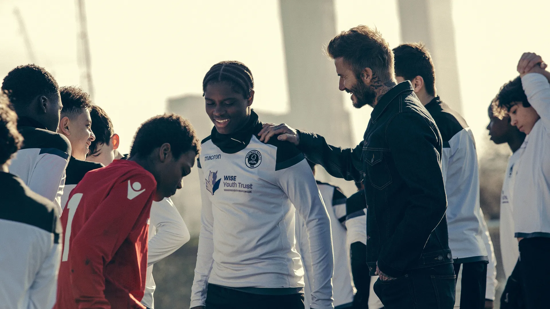 A sunny, outdoor scene from the sports docuseries Save Our Squad with David Beckham showing David Beckham in a denim jacket smiling and resting his hand on the shoulder of a smiling youth soccer player wearing a white Wise Youth Trust jersey on a training field.
