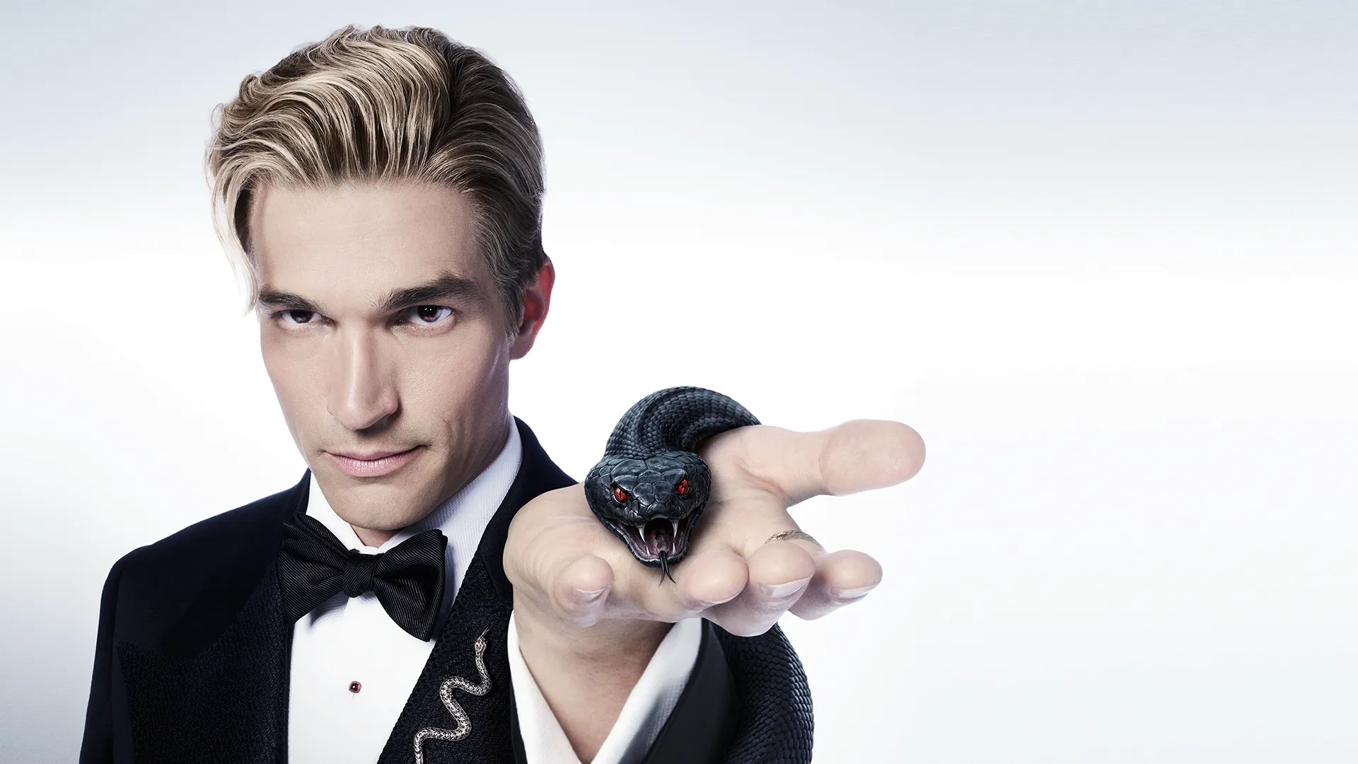 A man in a black tuxedo with slicked-back blonde hair holds out his open palm, revealing a coiled black snake with glowing red eyes and an open mouth.