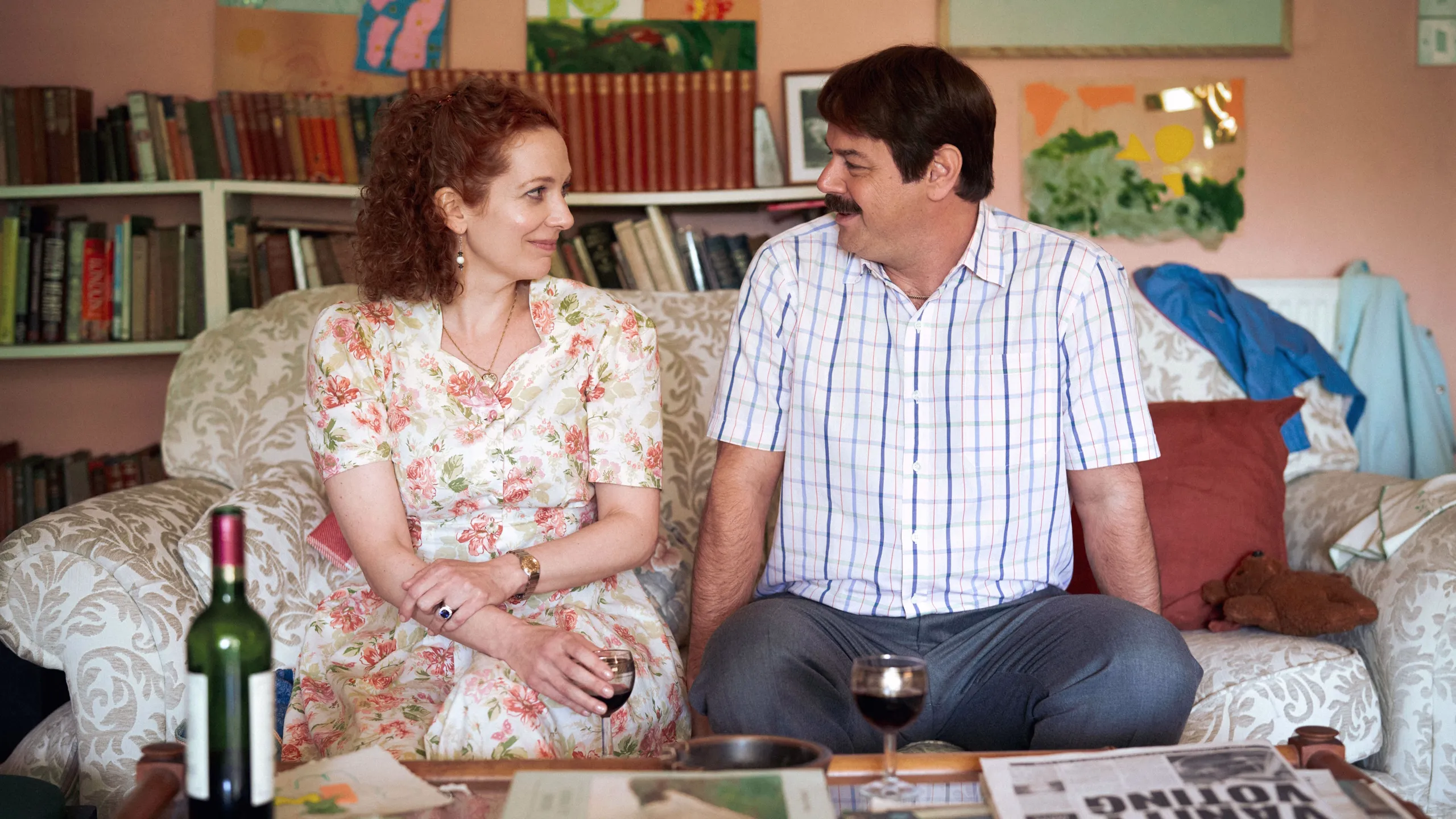 Lizzie Vereker (Katherine Parkinson) and Freddie Jones (Danny Dyer) sit together on a sofa with wine glasses in Rivals season 2.